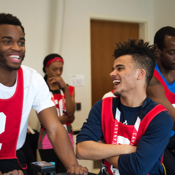 Two young men wearing red numbered pinnies laugh together while participating in an indoor cycling activity. In the background, other participants in similar attire are engaged in activities, creating a lively and energetic atmosphere.