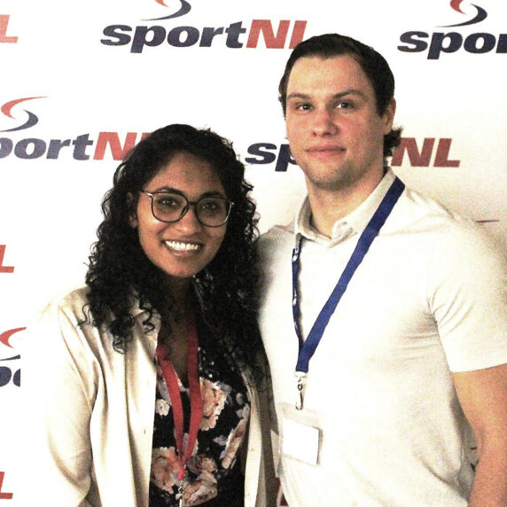 Two individuals smiling and posing in front of a SportNL backdrop. The woman on the left is wearing glasses, a floral top, and a light-colored blazer, while the man on the right is wearing a white collared shirt with a blue lanyard.