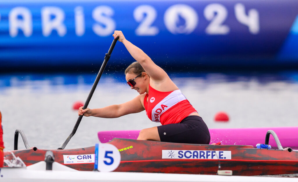 Canadian Paralympian Erica Scarff competes in Para canoe at the Paris 2024 Paralympic Games. She is wearing a red and white Team Canada uniform and paddles powerfully in her red and black boat, marked with her last name, "Scarff E." The race number "5" is visible on her boat. A large "Paris 2024" banner is seen in the blurred background.