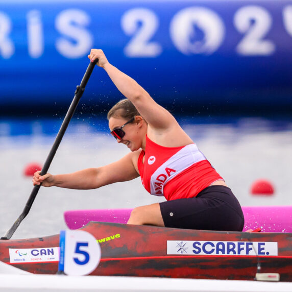 Canadian Paralympian Erica Scarff competes in Para canoe at the Paris 2024 Paralympic Games. She is wearing a red and white Team Canada uniform and paddles powerfully in her red and black boat, marked with her last name, "Scarff E." The race number "5" is visible on her boat. A large "Paris 2024" banner is seen in the blurred background.