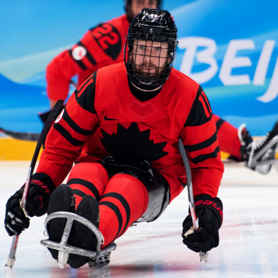 A Canadian Paralympic Team Para ice hockey player, wearing a red and black jersey with a black maple leaf emblem, competes at the Beijing 2022 Paralympic Winter Games. He is seated in a sled, gripping two hockey sticks with metal picks for propulsion. Another Canadian player is visible in the background on the ice, with the word "Beijing" displayed on the blue arena boards.