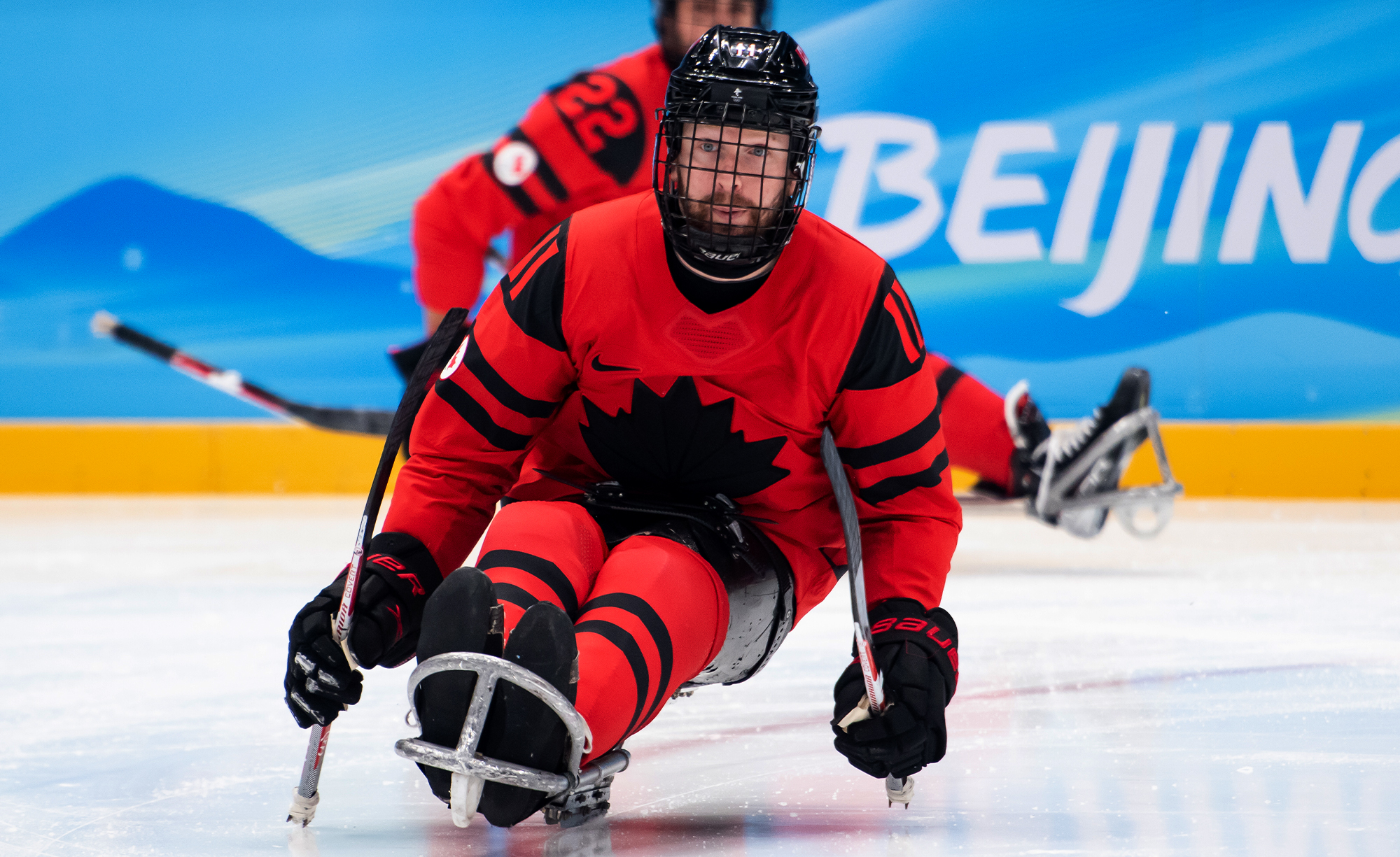 A Canadian Paralympic Team Para ice hockey player, wearing a red and black jersey with a black maple leaf emblem, competes at the Beijing 2022 Paralympic Winter Games. He is seated in a sled, gripping two hockey sticks with metal picks for propulsion. Another Canadian player is visible in the background on the ice, with the word "Beijing" displayed on the blue arena boards.