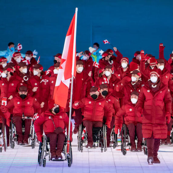 Canadian Paralympic Team athletes, dressed in matching red jackets and toques, proudly enter the stadium during the Opening Ceremony of the Paralympic Games. Some athletes wave Canadian flags, while others roll in wheelchairs. A large Canadian flag is carried at the front of the group. All athletes are wearing white face masks, and the background features blue lighting and a large red lantern.