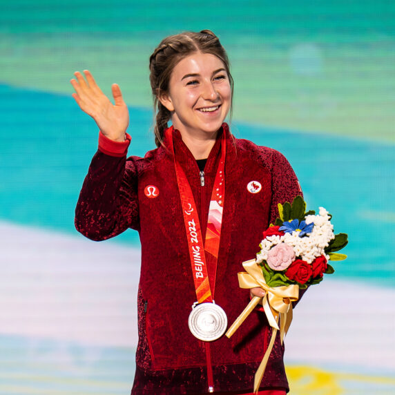 Canadian Paralympian Mollie Jepsen stands on the podium at the Beijing 2022 Paralympic Winter Games, wearing a red Team Canada jacket. She waves and smiles while holding her silver medal and a bouquet of flowers, celebrating her achievement in Para alpine skiing.