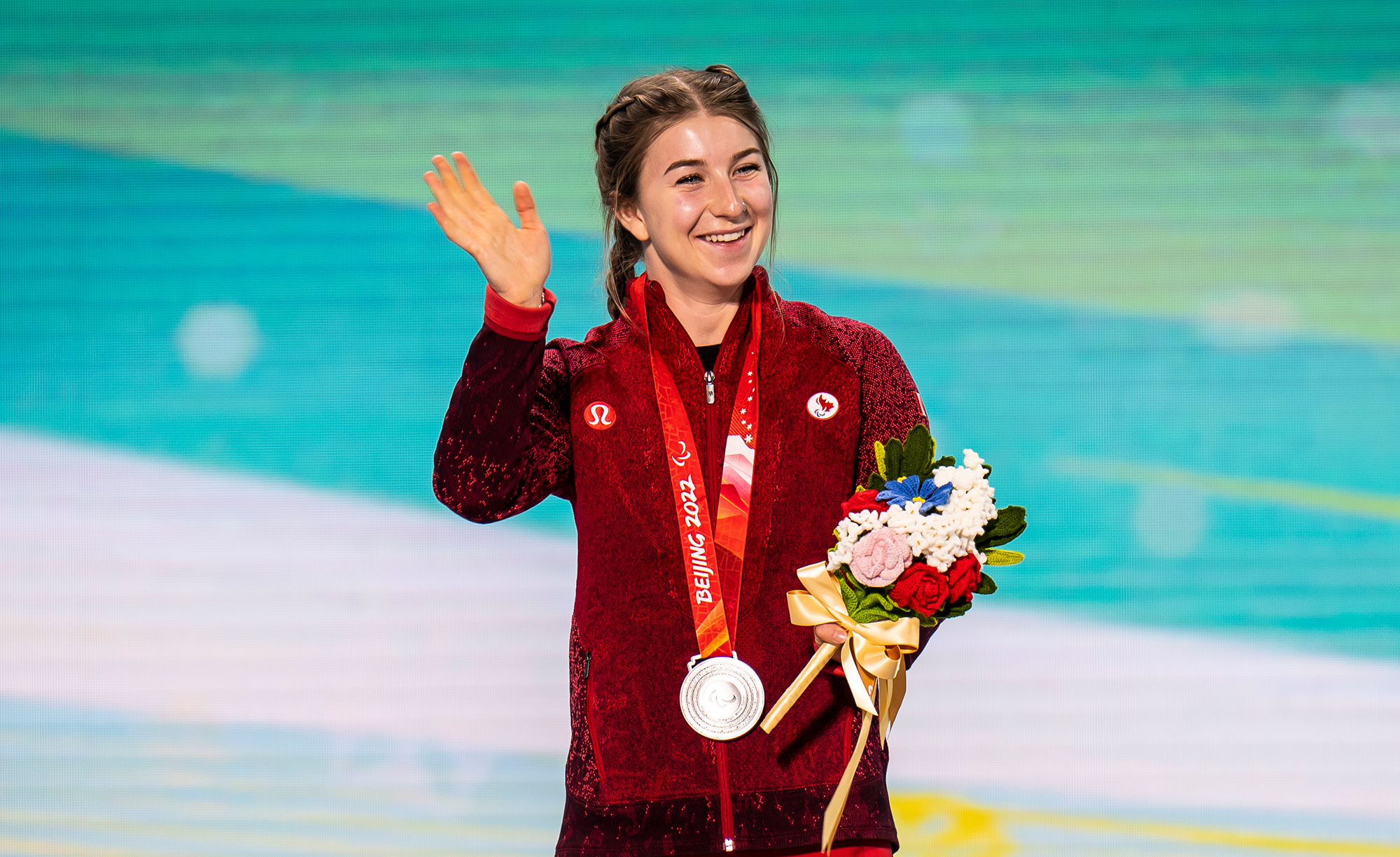 Canadian Paralympian Mollie Jepsen stands on the podium at the Beijing 2022 Paralympic Winter Games, wearing a red Team Canada jacket. She waves and smiles while holding her silver medal and a bouquet of flowers, celebrating her achievement in Para alpine skiing.