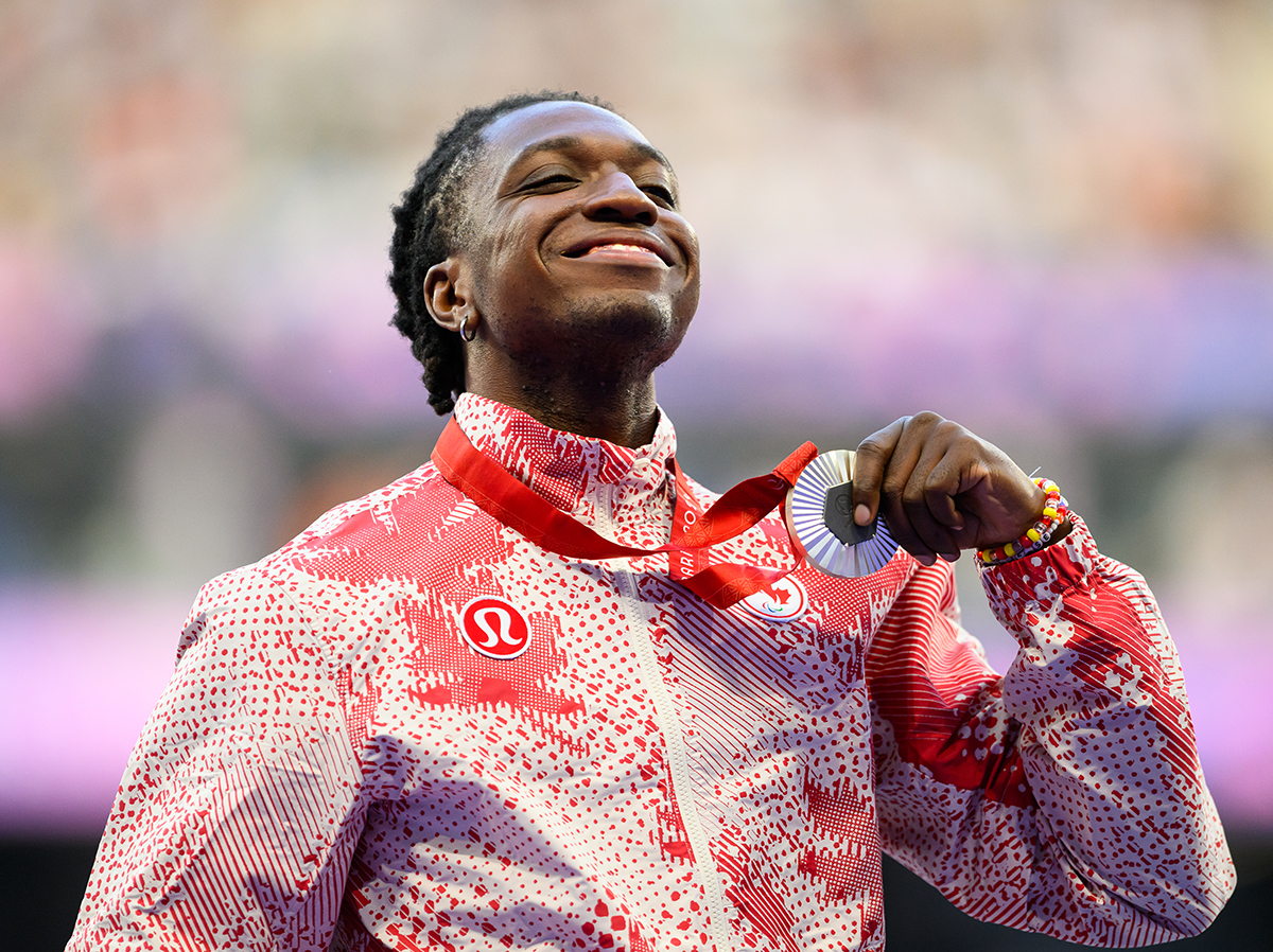 Canadian Paralympian Jesse Zesseu smiles proudly while holding up his silver medal at the Paris 2024 Paralympic Games. He is wearing Team Canada’s red and white lululemon podium jacket, featuring a bold abstract pattern. The blurred stadium lights in the background highlight the celebratory moment.