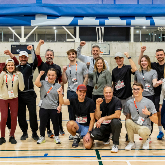 A group of participants at the Montreal ParaTough Cup 2024 pose together in a gymnasium, smiling and raising their fists in celebration. They are wearing athletic clothing and event lanyards, standing on a wooden sports court with a basketball hoop and blue banners visible in the background.