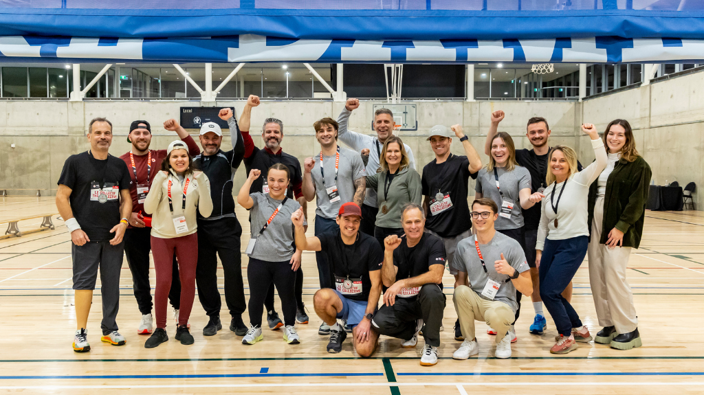 A group of participants at the Montreal ParaTough Cup 2024 pose together in a gymnasium, smiling and raising their fists in celebration. They are wearing athletic clothing and event lanyards, standing on a wooden sports court with a basketball hoop and blue banners visible in the background.