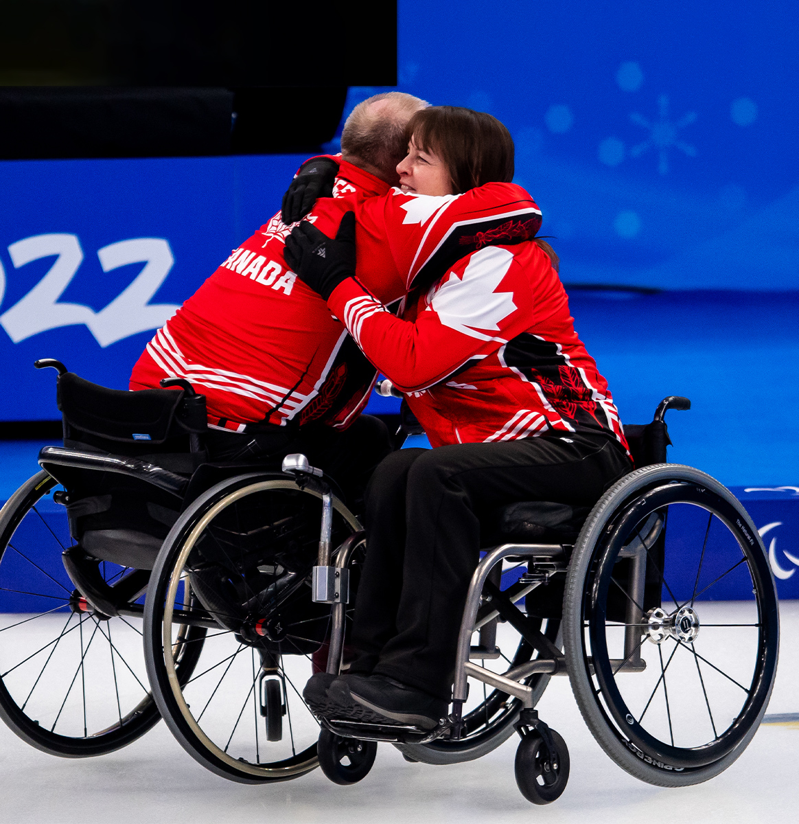 Two Canadian wheelchair curlers embrace in celebration on the ice. They are wearing matching red and white Team Canada jackets with maple leaf designs. Both athletes are in wheelchairs, and the background features a blue rink with the numbers "2022" visible, indicating the Beijing 2022 Paralympic Winter Games.