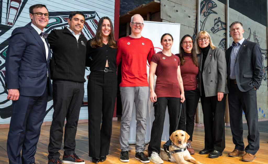 A group of eight people, including Canadian Paralympians, representatives, and a guide dog, pose together in front of a traditional Indigenous art backdrop. They are dressed in a mix of formal and casual attire, with some wearing red Team Canada apparel. The setting appears to be a special event or announcement related to the Canadian Paralympic Movement.