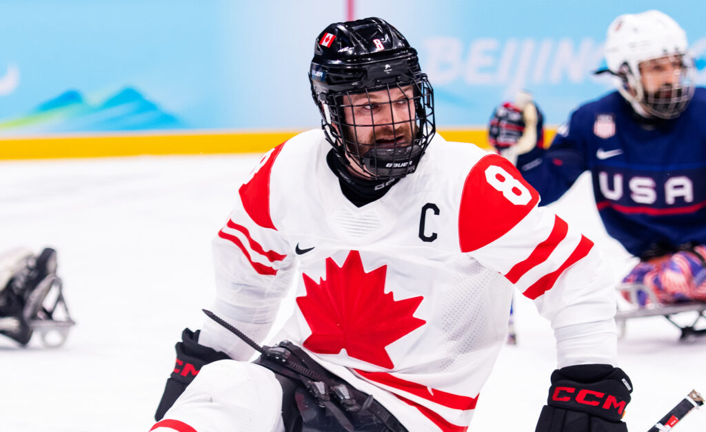 Canadian Para ice hockey player Tyler McGregor, wearing the captain’s "C" on his white Team Canada jersey with a red maple leaf, competes during a game. He is focused on the action, gripping his hockey sticks as a USA player is visible in the background.