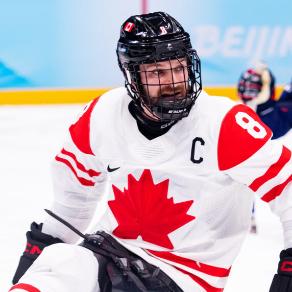 Canadian Para ice hockey player Tyler McGregor, wearing the captain’s "C" on his white Team Canada jersey with a red maple leaf, competes during a game. He is focused on the action, gripping his hockey sticks as a USA player is visible in the background.