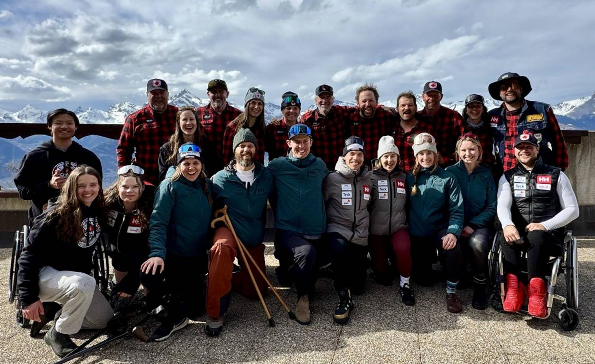 Group photo of the Canadian Para Alpine Ski Team smiling outdoors with snow-covered mountains in the background. Team members wear a mix of plaid jackets, alpine gear, and branded outerwear.