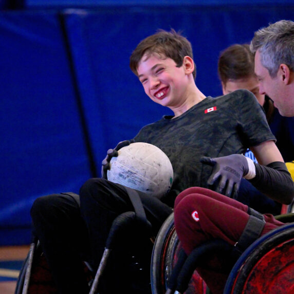 A young Canadian wheelchair rugby player smiles with joy while holding a rugby ball during a friendly match against Canadian Paralympian Byron Green. Both athletes are seated in sport wheelchairs on a gym court, sharing a moment of connection and fun.