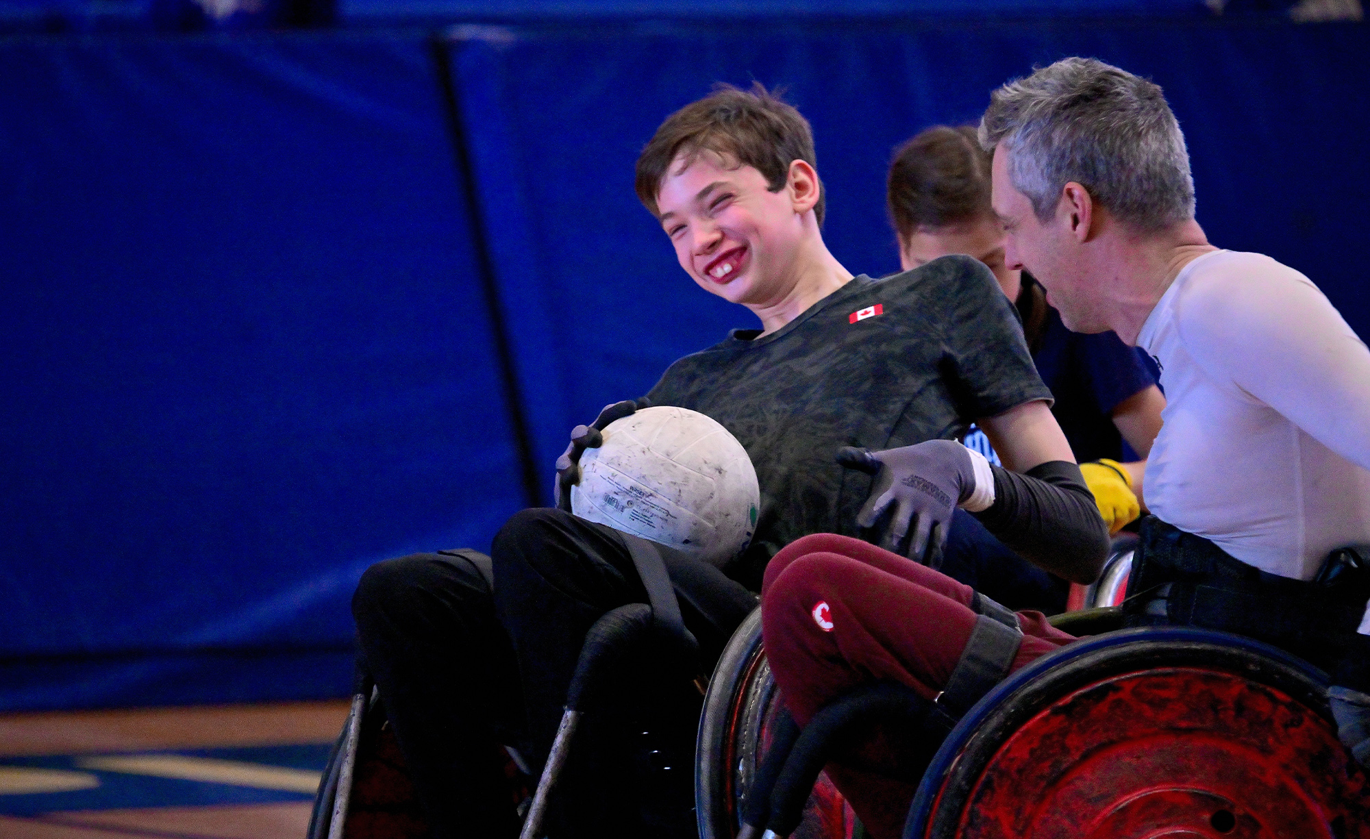 A young Canadian wheelchair rugby player smiles with joy while holding a rugby ball during a friendly match against Canadian Paralympian Byron Green. Both athletes are seated in sport wheelchairs on a gym court, sharing a moment of connection and fun.