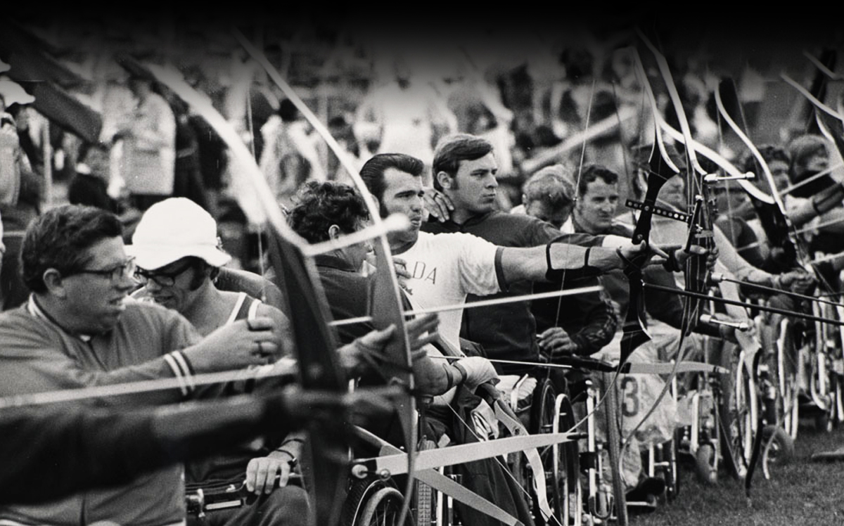 A black-and-white photo from the Heidelberg 1972 Paralympic Games captures a row of Para archers in wheelchairs, drawing their bows in competition. In the middle of the image, Canadian Paralympian Eugene Reimer wears a white shirt with "Canada" across the chest, focusing intently as he aims his bow. The background features a crowd of spectators watching the competition.