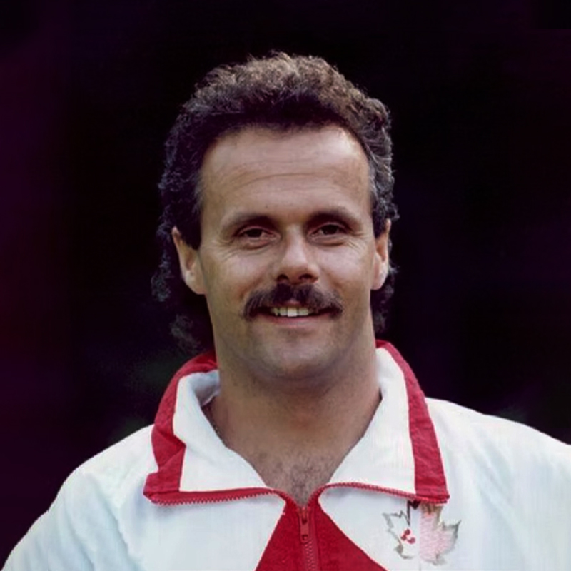 A close-up portrait of Hal Merrill, smiling in a white and red Team Canada tracksuit with a maple leaf emblem on the chest. The background is blurred, emphasizing his warm expression and signature mustache.