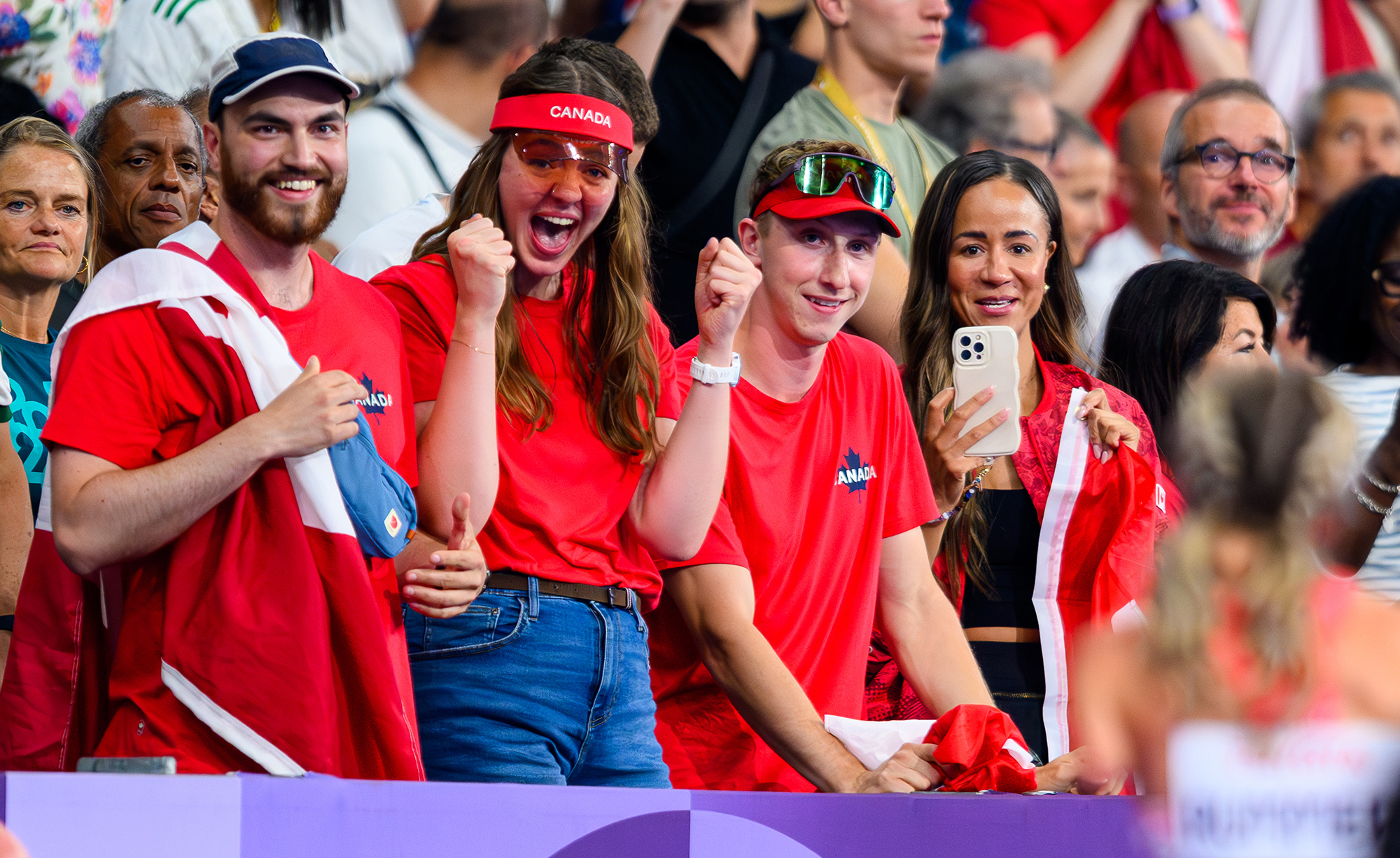 A group of enthusiastic Canadian fans, dressed in red and waving Canadian flags, cheer loudly in the stands for Canadian Paralympian Amanda Rummery at the Paris 2024 Paralympic Games. One fan captures the moment on a phone, while others raise their fists in excitement, showing their pride and support.