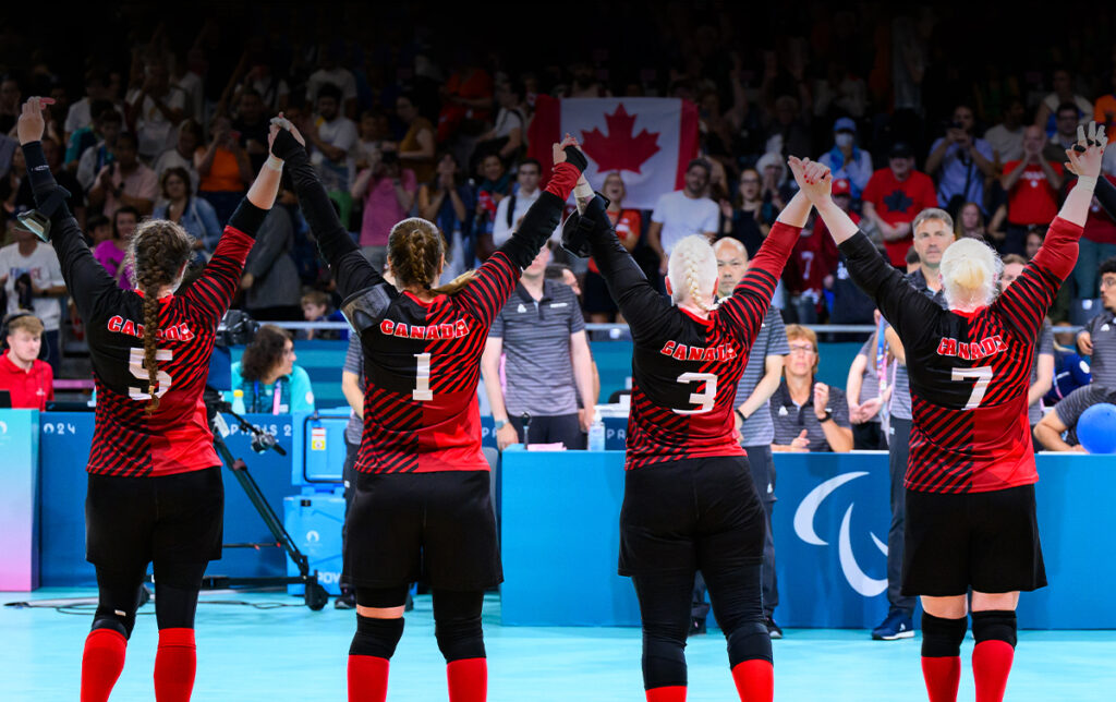The Canadian Women’s Goalball Team stands united with arms raised in celebration, facing the crowd after a match at the Paris 2024 Paralympic Games. Fans cheer and wave a Canadian flag in the background.
