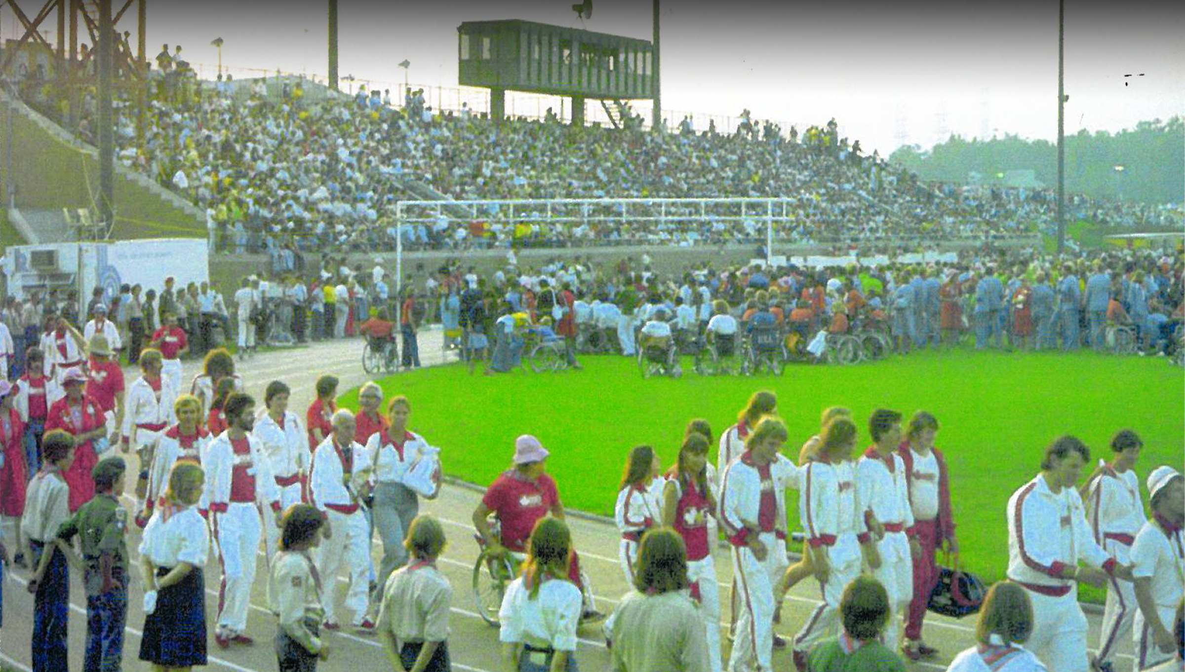 Paralympic athletes parade around a stadium track during the Opening Ceremony of the Toronto 1976 Paralympic Games, as a large crowd of spectators fills the stands.
