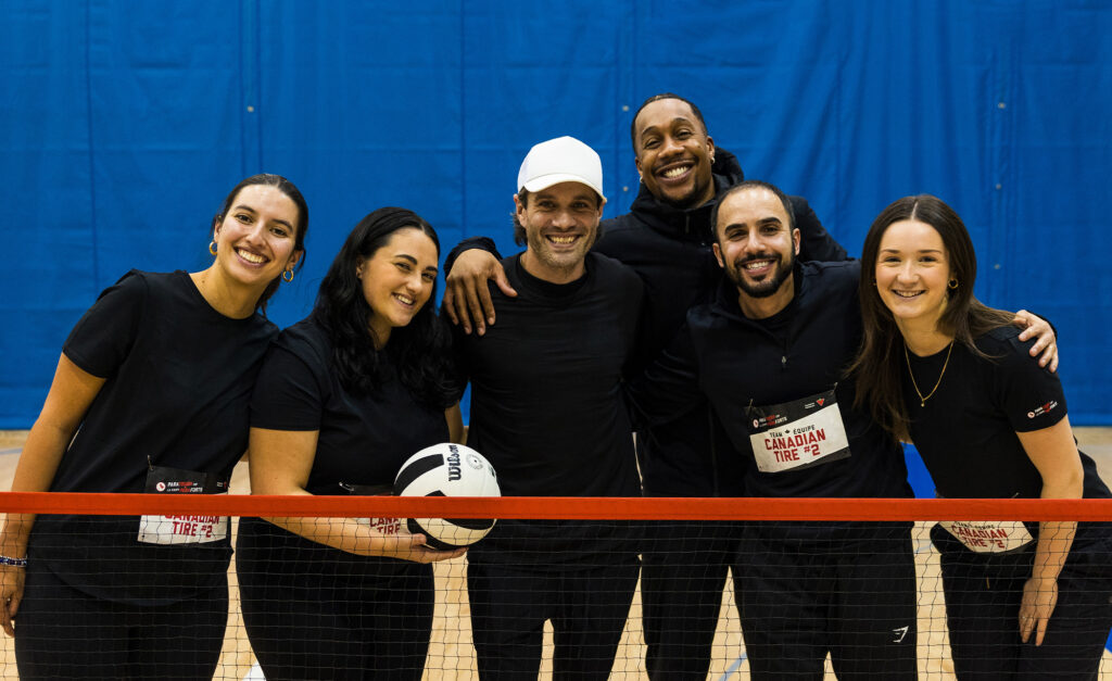 A smiling team from Canadian Tire poses together on a gym court behind a net at the ParaTough Cup Toronto event, holding a volleyball and wearing matching black shirts.