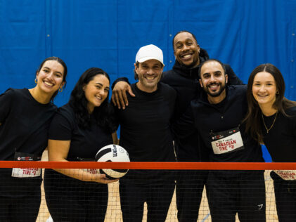 A smiling team from Canadian Tire poses together on a gym court behind a net at the ParaTough Cup Toronto event, holding a volleyball and wearing matching black shirts.