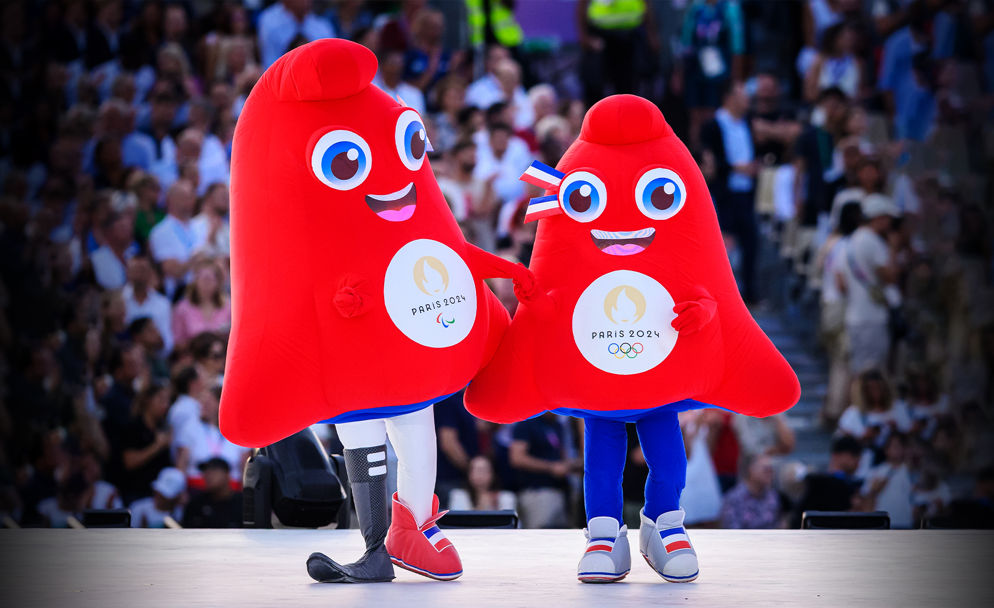 The Paris 2024 mascots, known as the Phryges, appear on stage during a public event. The red, hat-shaped characters are smiling and holding hands, one representing the Paralympics and the other the Olympics, each with the Paris 2024 emblem on their chests.