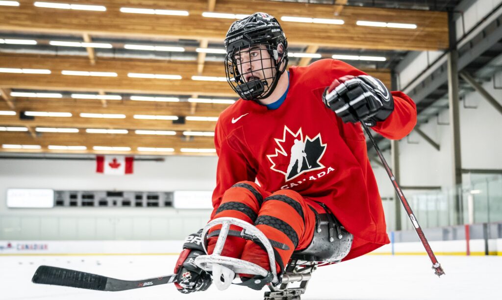 A Canadian Para hockey player in a red jersey with a maple leaf logo glides across the ice during practice, focused and mid-stride with a stick in hand at an indoor rink.