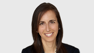 Dominique Breton, directrice au Comité paralympique canadien, sourit en portant un veston foncé et un collier de perles, sur un fond gris pâle.