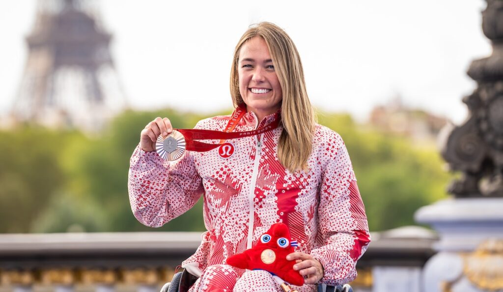 Canadian Paralympian Leanne Taylor smiles while holding her Paris 2024 bronze medal and Paralympic mascot plush toy. She wears a red and white Team Canada podium jacket, with the Eiffel Tower blurred in the background.