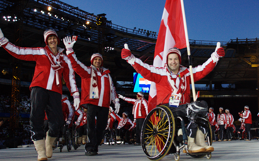 Canadian Paralympian Todd Nicholson leads Team Canada into the stadium at the Torino 2006 Paralympic Winter Games Opening Ceremony, proudly carrying the Canadian flag. He smiles and gives a thumbs-up while wearing the official red and white team uniform and a Canada tuque, surrounded by cheering teammates.