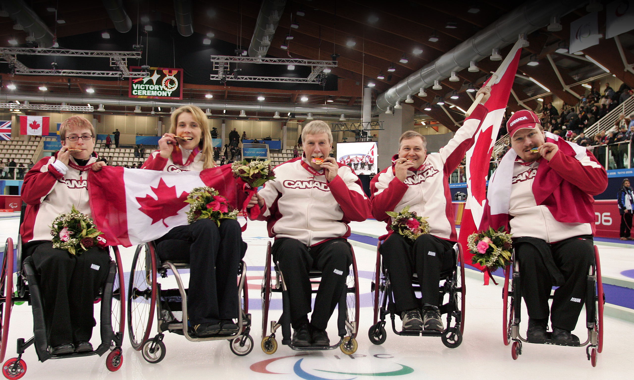 The Canadian Wheelchair Curling Team celebrates on the ice after winning gold at the Torino 2006 Paralympic Winter Games. The five athletes smile while holding bouquets and biting their gold medals, with two athletes holding Canadian flags.