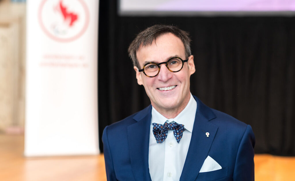 Marc-André Fabien, President of the Canadian Paralympic Committee, smiles while wearing a navy blue suit, patterned bow tie, and glasses at a formal event.