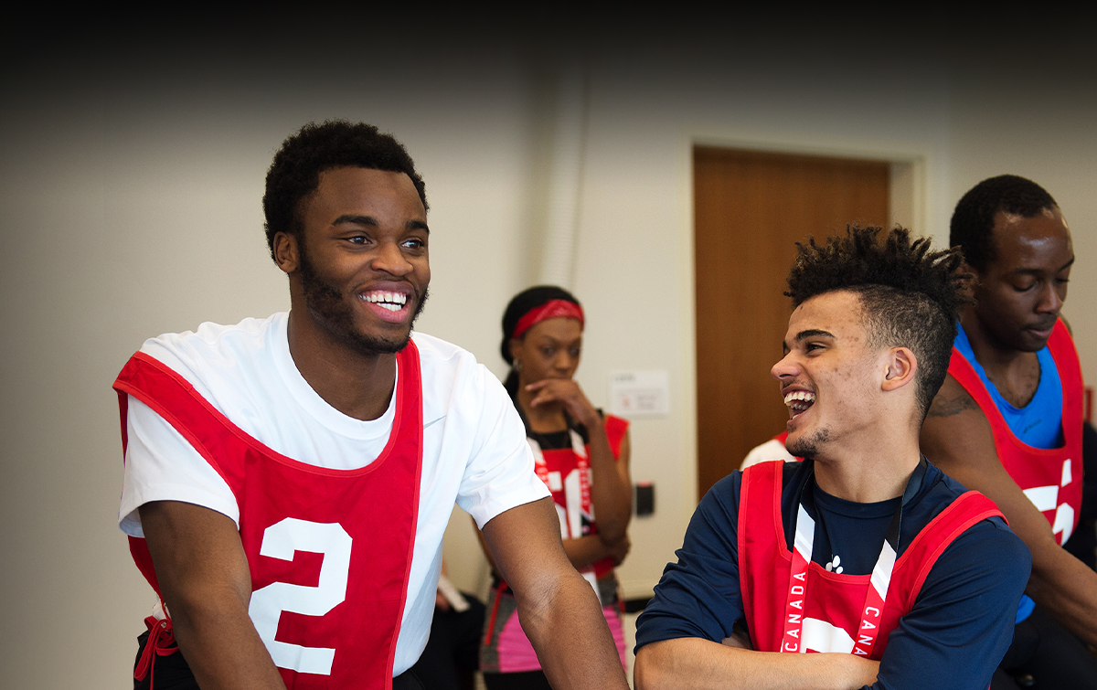 Two young men wearing red bibs laugh together during a stationary bike test at the 2016 Toronto Paralympian Search event, with other participants visible in the background.