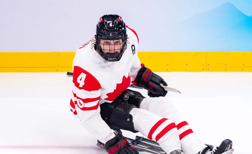 Canadian Para ice hockey player James Dunn wears jersey number 4 as he competes on the ice, focused and in motion, during a game. He wears a white Team Canada uniform with red accents and a black helmet.