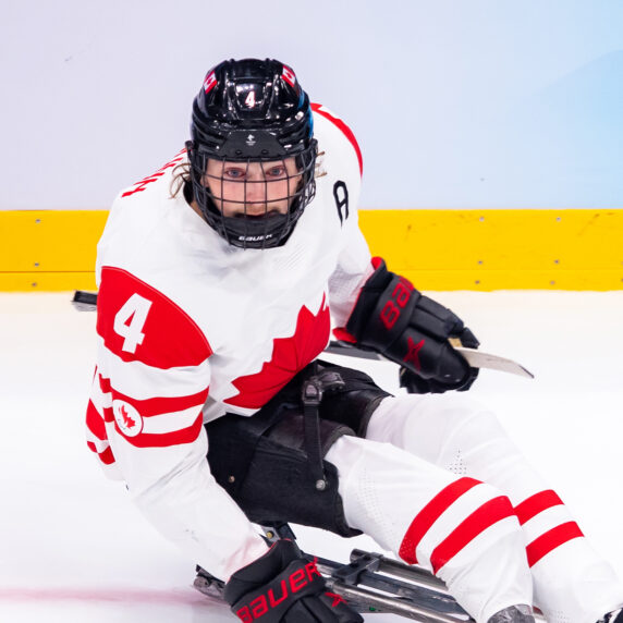 Canadian Para ice hockey player James Dunn wears jersey number 4 as he competes on the ice, focused and in motion, during a game. He wears a white Team Canada uniform with red accents and a black helmet.