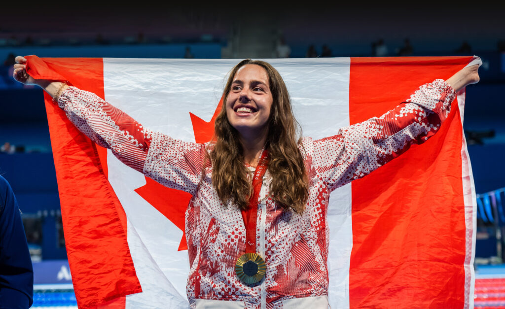 Canadian Paralympic swimmer Aurélie Rivard smiles proudly while holding a large Canadian flag behind her. She wears her Paris 2024 medal and Team Canada podium jacket on the pool deck.