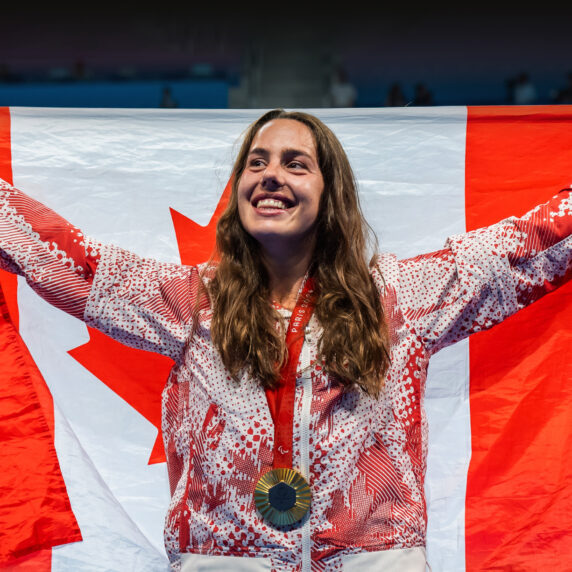 Canadian Paralympic swimmer Aurélie Rivard smiles proudly while holding a large Canadian flag behind her. She wears her Paris 2024 medal and Team Canada podium jacket on the pool deck.