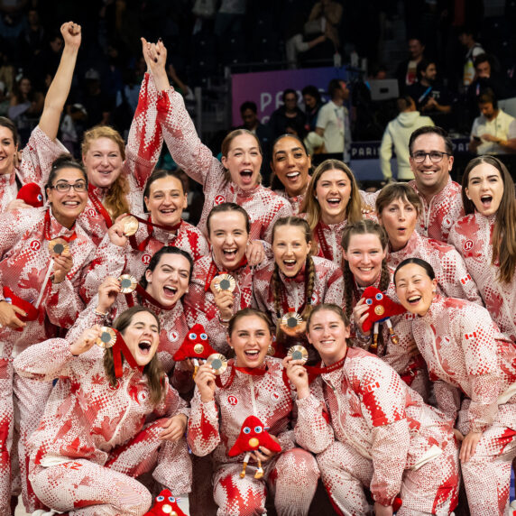 The Canadian women’s sitting volleyball team celebrates together in matching red and white Paralympic uniforms, proudly holding up their Paris 2024 bronze medals and mascots, smiling and cheering as a team.