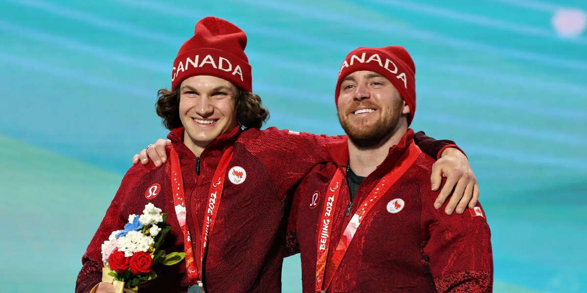 Canadian Paralympian Mac Marcoux and his guide stand together smiling on the podium at the Beijing 2022 Paralympic Winter Games, wearing matching red Team Canada toques and jackets with their medals around their necks.