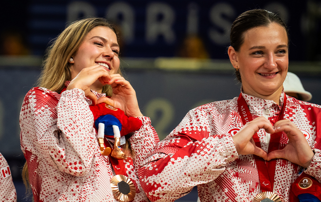 Two Canadian sitting volleyball players wearing Team Canada’s Paris 2024 uniforms and bronze medals smile and form heart shapes with their hands during the medal ceremony. One athlete holds the official Paralympic mascot.