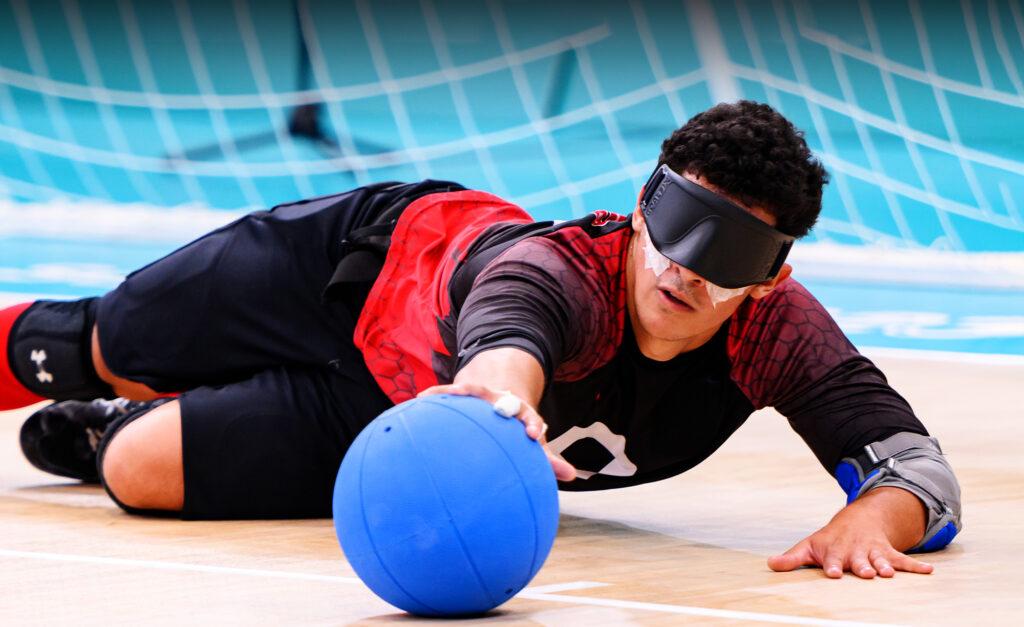 A Canadian goalball player dives to block a ball during competition at the Santiago 2023 Parapan American Games, wearing an eye mask and red-and-black team uniform on a light blue court.