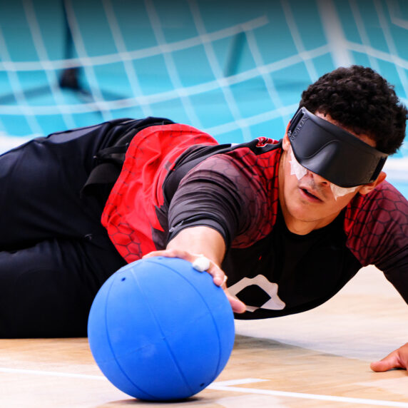 A Canadian goalball player dives to block a ball during competition at the Santiago 2023 Parapan American Games, wearing an eye mask and red-and-black team uniform on a light blue court.