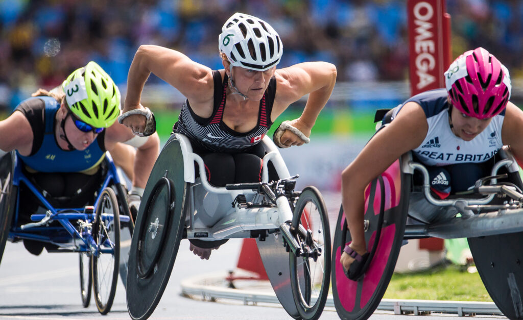 Canadian Paralympian Diane Roy races fiercely in her wheelchair on the track during a competition, positioned between athletes from Sweden and Great Britain, showing intense focus and power.