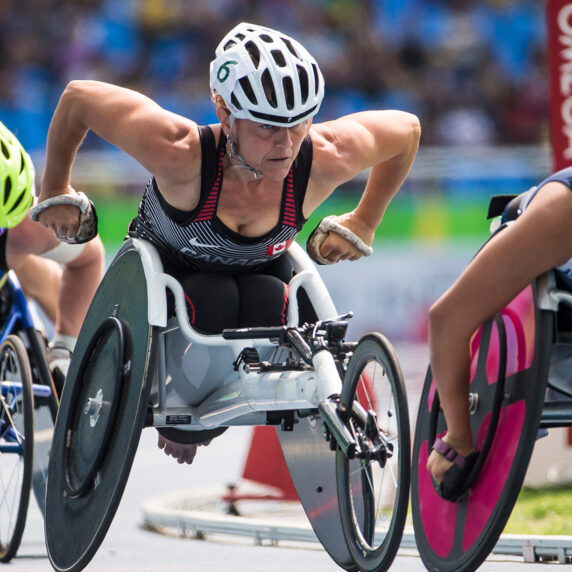 Canadian Paralympian Diane Roy races fiercely in her wheelchair on the track during a competition, positioned between athletes from Sweden and Great Britain, showing intense focus and power.