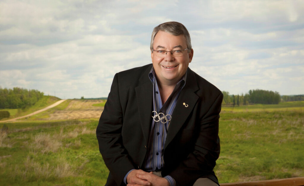 Dr. Robert Steadward smiles while seated in front of a scenic rural landscape. He wears a dark blazer, a blue-striped shirt, and a necklace featuring the Olympic rings, symbolizing his contribution to global sport and the Paralympic Movement.