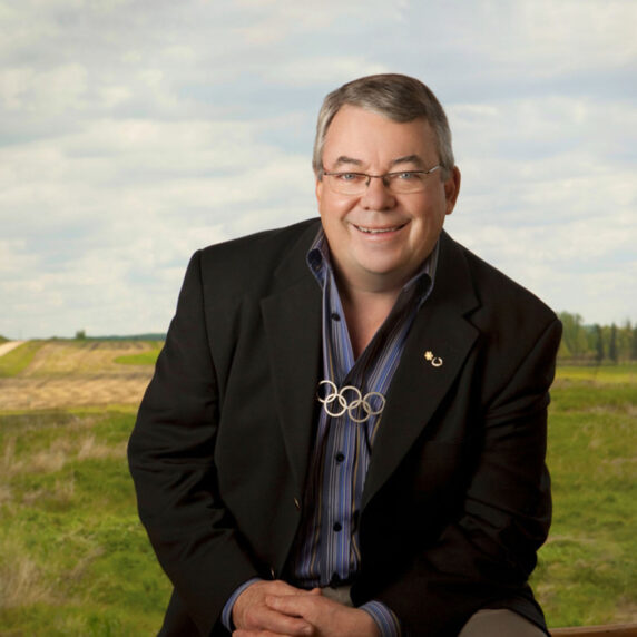 Dr. Robert Steadward smiles while seated in front of a scenic rural landscape. He wears a dark blazer, a blue-striped shirt, and a necklace featuring the Olympic rings, symbolizing his contribution to global sport and the Paralympic Movement.