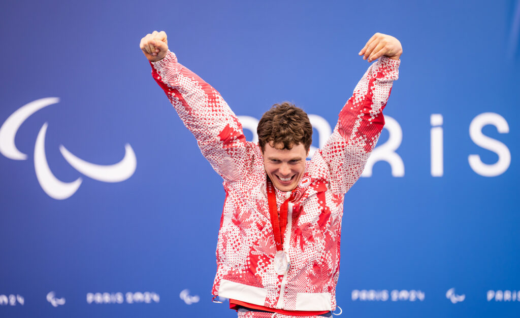Canadian Para swimmer Nathan Clement celebrates with both arms raised on the podium after winning a silver medal at the Paris 2024 Paralympic Games. He wears a red-and-white Team Canada uniform and smiles proudly in front of a blue backdrop featuring the Paralympic symbol and the word "Paris."