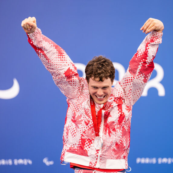 Canadian Para swimmer Nathan Clement celebrates with both arms raised on the podium after winning a silver medal at the Paris 2024 Paralympic Games. He wears a red-and-white Team Canada uniform and smiles proudly in front of a blue backdrop featuring the Paralympic symbol and the word "Paris."