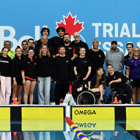 Group photo of the Para swimming squad and staff for Swimming Canada standing on the pool deck in front of a blue “Bell Trials” backdrop, with athletes and coaches smiling together.